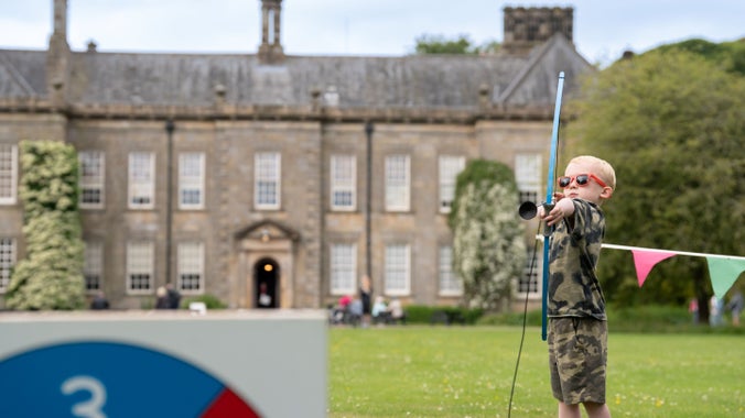 Young boy in khaki t-shirt and shorts and orange sunglasses is aiming a bow and arrow at a target with Wallington's house in the background. Part of Summer of Play, sponsored by Starling Bank.
